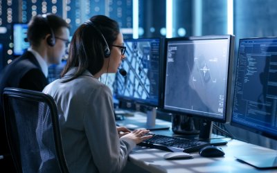 Female working in a Surveillance Team Gives Instructions with the Help of the Headsets. In the Background People Working and Monitors Show Various Information.